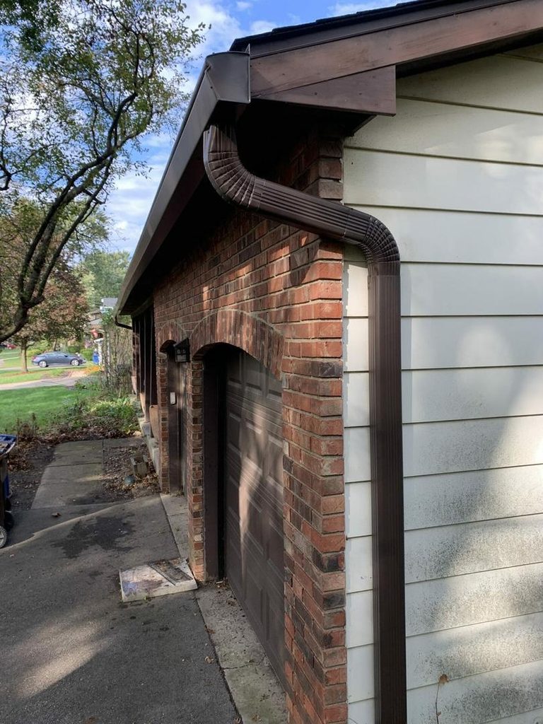 Brown downspout carrying water from a hooded gutter down a brick garage wall on a Central Ohio home